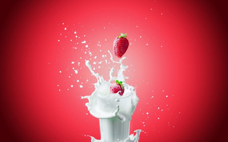 View of nice fresh red strawberry falling down in to the glass milk making a big splash on a red background